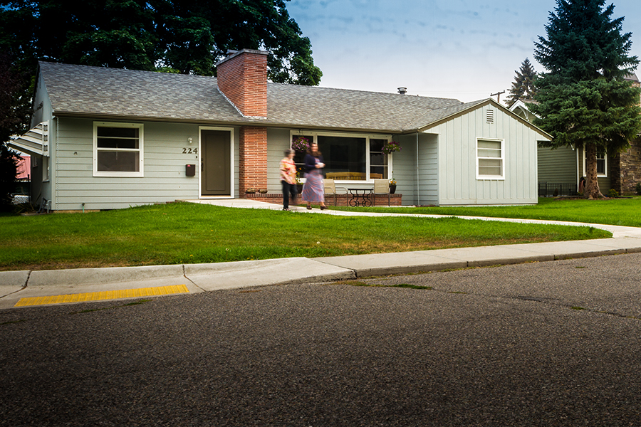 Two women continue walking along a sidewalk away from a one-story house. The path goes through a grassy lawn and connects to a passing sidewalk, which has a curb-ramp that leads to the street.